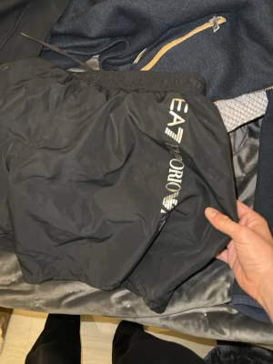 Svarta shorts från Emporio Armani EA7 - Säljer ett par svarta shorts från Emporio Armani EA7 med resår i midjan och logga tryckt på sidan. Shortsen har en enkel och sportig design, perfekta för en avslappnad stil. Materialet känns lätt och syntetiskt.