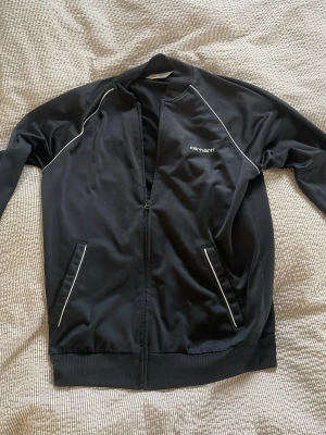 Svart trackertröja från Carhartt - Snygg svart trackietröja från Carhartt med vita detaljer längs sömmarna och dragkedja framtill. Jackan har ribbade muddar, två fickor och en klassisk sportig look. Perfekt för dig som gillar streetwear och enkel stil.