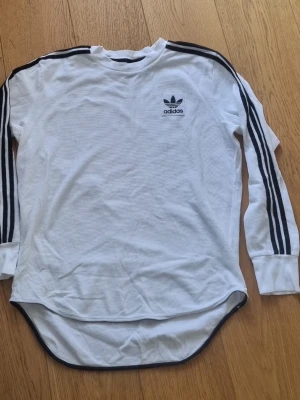 Vit långärmad Adidas tröja - Säljer en vit långärmad tröja från Adidas med svarta klassiska ränder längs ärmarna och Adidas-logga på bröstet. Tröjan har rund halsringning, ribbade muddar och längre rundad nederkant. Perfekt för en sportig och avslappnad stil.