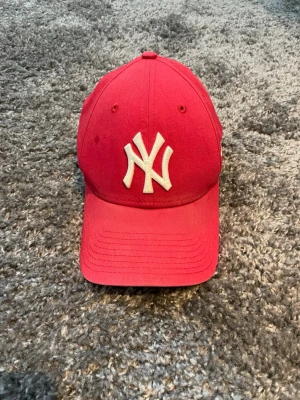 Röd New Era Yankees keps 9FORTY - Säljer en röd New Era 9FORTY keps med klassisk böjd skärm och justerbar rem bak. Framtill sitter ett vitt broderat New York Yankees-logo. Kepsen är tillverkad i bomull och har ventilerande hål upptill. Perfekt för dig som vill ha en sportig och stilren look.