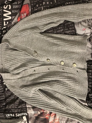 Ljusgrå ribbad kofta med knappar - Snygg ljusgrå ribbad kofta med v-ringning och fyra ljusa knappar framtill. Koftan har långa ärmar och är tillverkad i ett mjukt, stickat material som känns skönt mot huden. Perfekt att slänga över en t-shirt för en avslappnad look.