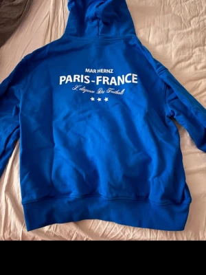 Blå hoodie från Mar Hernz Paris-France - Snygg blå hoodie från Mar Hernz med tryck 'PARIS-FRANCE' och textdetaljer på ryggen samt liten logga fram. Klassisk känguruficka och huva. Perfekt för dig som gillar streetwear och vill sticka ut med en unik design.