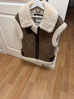 Brun pälsväst från Zara med dragkedja - Snygg brun pälsväst från Zara med bred krage i vit fuskpäls och detaljer i svart. Västen har dragkedja framtill, två fickor och dekorativa spännen vid sidorna. Perfekt för lager på lager och ger en cool look till din outfit. Nypris 979kr säljes kanonpris 449kr 