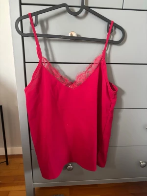 Röd linnetopp med spetskant - Snygg röd linnetopp med tunna axelband och spetsdetalj längs v-ringningen. Perfekt att styla till jeans eller kjol för en trendig look. Luftigt material och enkel design gör den superenkel att matcha.