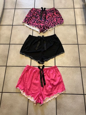 Tre pyjamasshorts med rosett storlek M - Säljer tre pyjamasshorts i olika stilar: ett rosa med leopardmönster och vit volangkant, ett svart med vit volangkant och ett enfärgat rosa med vit volangkant. Alla har en svart rosett framtill och elastisk midja. Perfekta för mysiga kvällar.