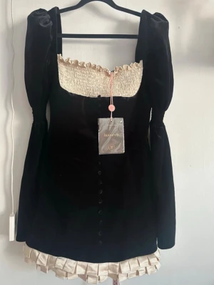 Svart sammetsklänning House of CB Azaria dress  - Jättefin svart sammetsklänning från House of CB med puffiga långärmar, krämfärgad smockad överdel och volangkant nertill. Klänningen har dekorativa knappar framtill och dragkedja i ryggen. Regular cup size/regular length. Liten i storlek! ✨Säljer den då jag tyvärr inte kan ha den då den är för liten för mig! Annonsen ligger även ute på vinted (@/lasangeplattan) Nypris 2700kr