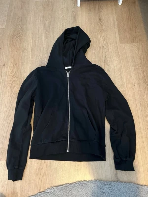 Svart hoodie med dragkedja från 157 - Klassisk svart hoodie från 157 med dragkedja framtill och två fickor. Tröjan har huva och ribbade muddar vid ärmslut och nederkant. Perfekt för en avslappnad stil och enkel att matcha med det mesta. Mjuk insida för extra komfort.
