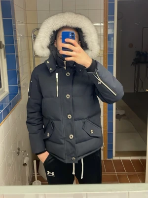 Mörkblå dunjacka med pälskant - Säljer en mörkblå dunjacka från Moose Knuckles med stor huva och vit pälskant. Jackan har flera dragkedjefickor, stora knappar och detaljer i silver. Perfekt för kalla vinterdagar och riktigt snygg streetstil.