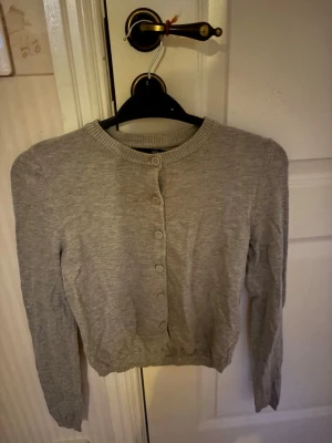 Ljusgrå kofta med knappar - En enkel och stilren ljusgrå kofta med rund halsringning och knappar framtill. Koftan har långa ärmar och ribbade muddar vid ärmslut och nederkant. Perfekt att slänga över en t-shirt eller linne för en avslappnad look.