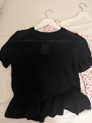 Svart stickad topp från Divided - Säljer en svart stickad topp från Divided med korta ärmar och volangkant nedtill. Tröjan har ett diskret hålmönster och ribbad halsringning. Perfekt till jeans eller kjol för en trendig look.