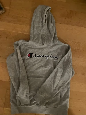 Grå hoodie från Champion - Säljer en grå hoodie från Champion med stor logga broderad på bröstet. Klassisk känguruficka framtill och mjukt material som känns skönt mot huden. Perfekt för dig som gillar sportig och avslappnad stil. Huva med dragsko för extra komfort.