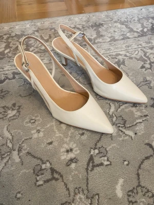 Vita slingback pumps med spetsig tå - Stilrena vita pumps med spetsig tå och öppen häl. Skorna har en smal klack och justerbar rem med silverfärgat spänne. Tillverkade i ett slätt material som ger en elegant look. Perfekta för att lyfta vilken outfit som helst.