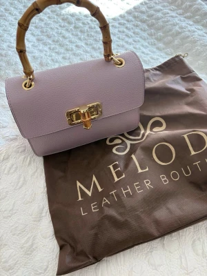 Oanvänd! Ljuslila handväska med bambuhandtag - Supersnygg ljuslila handväska från Melodi Leather Boutique i äkta läder. Väskan har ett unikt handtag i bambu, guldfärgade detaljer och kedjerem. Stängs med ett elegant guldfärgat spänne framtill. Perfekt accessoar för att lyfta din outfit.