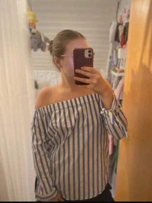 Randig offshoulder topp blå/vit - Säljer en snygg offshoulder topp med blå och vita vertikala ränder. Toppen har långa ärmar med resår vid axlarna och ärmarna, vilket ger en avslappnad och trendig look. Perfekt för dig som gillar en stilren och fräsch outfit.