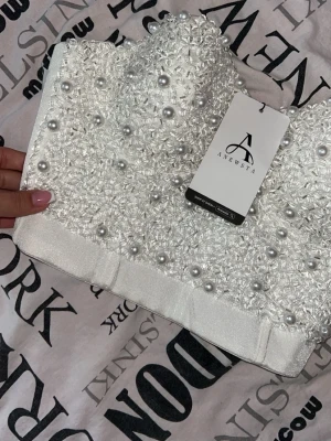 Vit pärlprydd korsett från Anewsta - Supersnygg vit korsettopp från Anewsta, täckt med glittriga pärlor och dekorativa detaljer. Toppen har en strukturerad form och är perfekt för att sticka ut. Materialet känns lyxigt och toppen är axelbandslös för en trendig look.