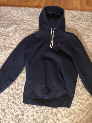 Mörkblå hoodie från Polo Ralph Lauren - Snygg mörkblå hoodie från Polo Ralph Lauren med klassisk huva och vita dragsnören. Har en känguruficka framtill och den ikoniska röda loggan broderad på bröstet. Perfekt för en chill och avslappnad stil.