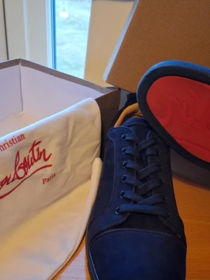 Mörkblå sneakers från Christian Louboutin - Snygga mörkblå sneakers från Christian Louboutin med klassisk röd sula och diskret logga. Skorna har rund tå, matchande mörkblå snörning och är tillverkade i mocka för en lyxig känsla. Perfekta för dig som vill sticka ut med stil.