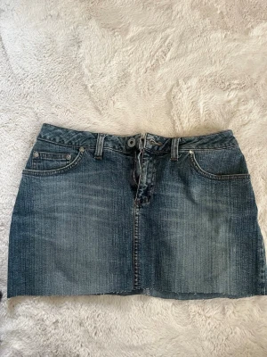 Blå jeanskjol från Levi's - Säljer en klassisk blå jeanskjol från Levi's med fem fickor, bälteshällor och knappgylf. Kjolen har en rak, kort passform och är tillverkad i slitstarkt denimtyg. Perfekt för dig som gillar en tidlös och avslappnad stil.