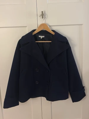 Mörkblå kappa från Design by Si - Stilren mörkblå rock från Design by Si med bred krage och dubbelknäppning framtill. Kappan har två sidofickor och uppvikta ärmslut för en modern look. Tillverkad i ett mjukt, tjockare tyg som passar perfekt till kyligare dagar.