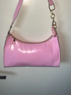 Rosa axelremsväska i lack - Snygg rosa handväska i glansigt lackmaterial med guldfärgade metalldetaljer och dragkedja. Väskan har en bred axelrem och svart innerfoder. Perfekt för dig som gillar en poppig och trendig accessoar.