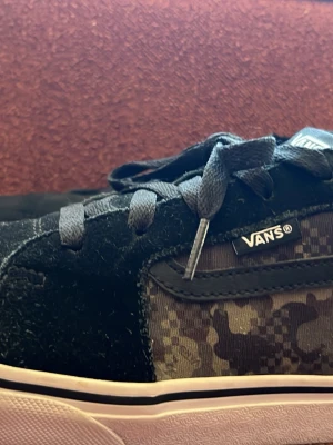 Svarta Vans sneakers med camomönster - Säljer ett par svarta Vans sneakers med coolt camomönster på sidan. Skorna har ovandel i mocka och textil, klassiska svarta skosnören och vit sula med svart rand. Perfekta för dig som gillar streetwear och vill ha något unikt på fötterna.