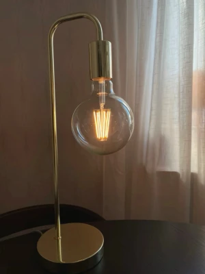 Två stilrena bordslampor  - Säljer en stilren bordslampa i mässingfärgad metall med rund fot och böjd arm. Lampan har en stor, rund glödlampa med synlig glödtråd som ger en modern och industriell känsla. Perfekt som inredningsdetalj för skrivbord eller nattduksbord.