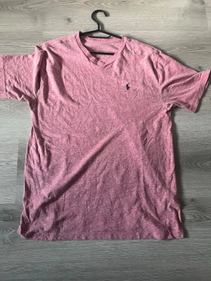 Rosa t-shirt från Ralph Lauren - Säljer en rosa t-shirt från Ralph Lauren med klassisk v-ringning och den ikoniska broderade loggan på bröstet. T-shirten är kortärmad och gjord i mjuk bomull, perfekt för en avslappnad stil.