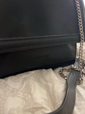Svart axelväska från Don Donna - Snygg svart axelväska från Don Donna i slätt konstläder. Väskan har en stilren design med silverfärgad kedjerem och flera fack inuti för smart förvaring. Perfekt för dig som gillar en klassisk och enkel look.