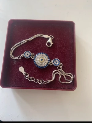 Silverarmband med blått och vitt öga - Snyggt armband i äkta silver (stämplat 925) med tre runda detaljer i blått, vitt och gult. Armbandet har ett klassiskt öga-motiv med gnistrande stenar och justerbar kedja. Perfekt accessoar för att lyfta din stil.