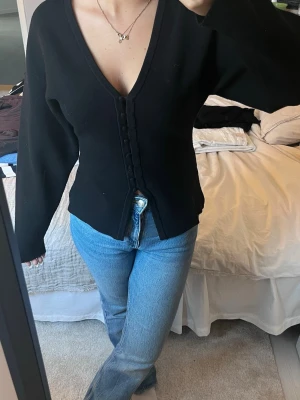 Svart kofta med knappar och v-ringning - Snygg svart kofta med djup v-ringning och dekorativa knappar framtill. Modellen är figurnära med långa ärmar och lätt utsvängd nederkant. Perfekt att styla med jeans eller kjol för en trendig look.