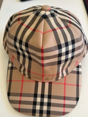 Beige Burberry keps med rutmönster - Säljer en klassisk beige keps från Burberry med det ikoniska rutmönstret i svart, vitt och rött. Kepsen har böjd skärm och justerbar rem bak med Burberry London England-märke. Tillverkad i bomull för en skön och stilren look.