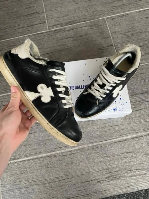 Svarta sneakers från Gallery Dept - Säljer ett par svarta sneakers från Gallery Dept med vit sula och vita detaljer, inklusive en stor vit klöver på sidan. Skorna har vita snören och mjuk, lurvig insida för extra komfort. Tillverkade i läder med en unik och trendig design.