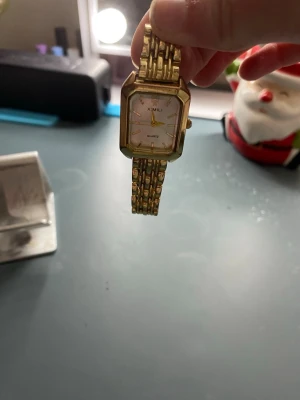 Guldfärgad klocka från XIMILI - Stilren guldfärgad klocka från XIMILI med rektangulärt ur och länkarmband i metall. Urtavlan är vit med guldiga detaljer och quartz-urverk. Perfekt accessoar för dig som gillar klassisk och elegant stil.