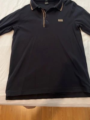 Svart långärmad polotopp från BOSS - Snygg svart långärmad polotopp från BOSS med gulddetaljer på kragen och logga på bröstet. Klassisk pikékrage och knappslå med BOSS-tryck. Perfekt för en stilren och clean look.