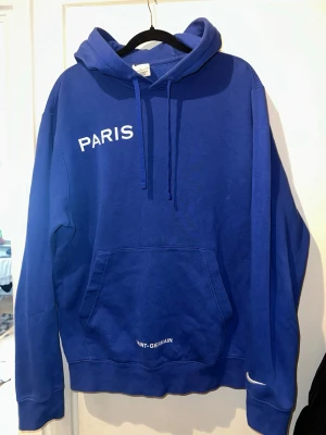 Blå Nike Paris Saint-Germain hoodie - Snygg blå hoodie från Nike med texten PARIS på bröstet och Saint-Germain nedtill. Klassisk känguruficka, huva med dragsko och Nike-logga på ärmen. Perfekt för dig som gillar sportig stil och vill representera PSG. Tillverkad i mjukt bomullsmaterial.