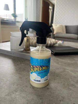 Lush Super Milk Glitter Mist - Super Milk Glitter Mist från Lush i en genomskinlig plastflaska med svart spraymunstycke. Innehållet är vitt och etiketten har en färgglad design med blå bakgrund och moln. Perfekt för dig som vill ha extra glans och doft i håret.