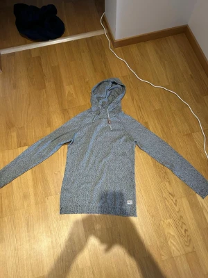 Gråmelerad hoodie med dragsko - Fast pris. Säljer en gråmelerad hoodie med huva och dragsko. Tröjan har lång ärm och är tillverkad i ett mjukt, stickat material som ger en avslappnad look. Liten etikett nertill på framsidan. Perfekt för dig som gillar enkel och stilren design. 