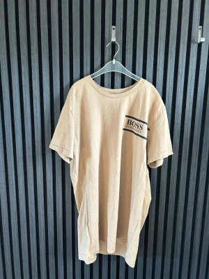 Beige t-shirt från Hugo Boss (använt 2-3 tillfällen) - Snygg beige t-shirt från Hugo Boss med klassisk rund hals och svart logga på bröstet. Tillverkad i mjuk bomull och har en avslappnad passform med korta ärmar. Perfekt för en clean och stilren look.