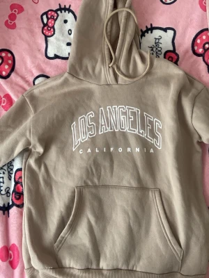 Beige hoodie från SHEIN med tryck - Snygg beige hoodie från SHEIN med stor vit text 'LOS ANGELES CALIFORNIA' på bröstet. Hoodien har huva med dragsko, magficka och ribbade muddar. Tillverkad i 100% polyester, perfekt för en chill och avslappnad stil. Knappt använd