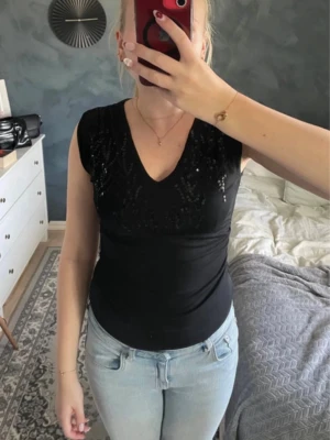 Svart v-ringad topp med paljetter - Snygg svart topp med v-ringning och paljettdetaljer framtill. Ärmlös modell som sitter tight och har en enkel, stilren rygg. Perfekt att styla med jeans eller kjol 