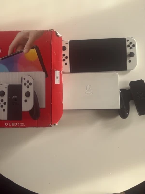 Nintendo Switch OLED vit konsol - Nintendo Switch OLED konsol i vit färg med matchande vita Joy-Con handkontroller och vit dockningsstation. Konsolen har en stor OLED-skärm och kan användas både bärbart och kopplad till TV. Originalkartong och ingen Joy-Con grip fast alla cabler finns