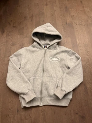 Grå zip hoodie från Corteiz - Säljer en grå zip hoodie från Corteiz med stor vit logga på ryggen och mindre tryck på bröstet. Hoodien har huva, fickor framtill och ribbade muddar vid ärmar och midja. Tillverkad i mjukt sweatshirtmaterial, perfekt för en chill och streetwear-inspirerad look.