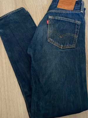 Levi's blå raka jeans - Säljer ett par klassiska blå jeans från Levi's i modellen 501 ’54. Jeansen har en mörk tvätt och detaljer i kontrastsömmar samt en brun läderpatch bak i midjan. Tillverkade i slitstark denim, perfekta till vardagslooken. Helt nya oanvända endast provade hemma några gånger. Stl w28 l32. Hör av dig vid frågor! Pris kan diskuteras vid snabb affär!😊