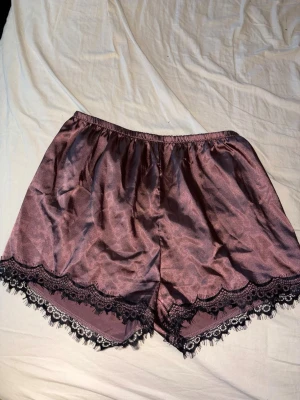 Rosa satinpyjamasshorts med spets - Snygga pyjamasshorts i glansigt rosa satinmaterial med svart spetskant nedtill. Shortsen har elastisk midja för skön passform och är tillverkade i 95% polyester och 5% elastan. Perfekta för mysiga kvällar hemma. storlek M