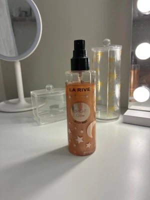 La Rive Bright Glow Body Mist - La Rive Bright Glow Body Mist i en genomskinlig plastflaska med svart spraymunstycke. Innehållet är skimrande kopparfärgat och flaskan har stjärnor och månar som dekoration. Perfekt för dig som vill ha en fräsch och glittrig body & hår mist