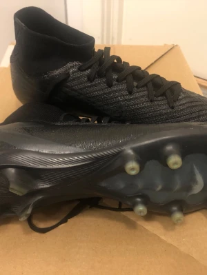 Svarta Superflys elit med strumpa - Säljer ett par helsvarta Nike superflys elit med strumpa och som har ag PRO dobbar. Skorna har en modern, strömlinjeformad design med snörning och ovandel i flyknit som ger bra bollkontroll. Den är bra för dig som kan springa fort vid kanten och perfekta för dig som vill ha stilrena och funktionella skor på planen.