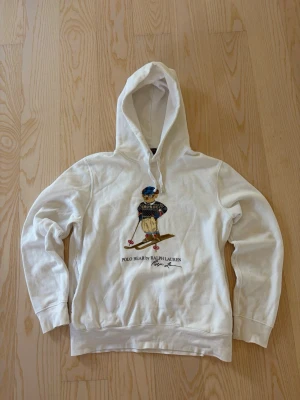 Vit hoodie Polo Bear Ralph Lauren - Vit hoodie från Ralph Lauren med den ikoniska Polo Bear-figuren på framsidan, klädd i vinterkläder och på skidor. Hoodien har huva med dragsko och ribbade muddar. Tillverkad i mjukt material som känns skönt mot huden. Perfekt för en avslappnad och cool stil.