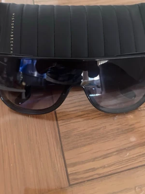 Svarta solglasögon Victoria Beckham - Snygga svarta solglasögon från Victoria Beckham med bred båge och mörka glas. Sidorna har tunna metallskalmar i silverfärg som ger en exklusiv känsla. Perfekt accessoar för att lyfta din stil i solen.