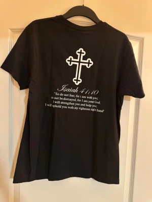 Svart t-shirt med kors och citat - Svart t-shirt från Next Level med vitt kors-tryck på bröstet och större kors samt bibelcitat på ryggen. Klassisk rund hals och korta ärmar. Tillverkad i mjuk bomull som känns skön mot huden.