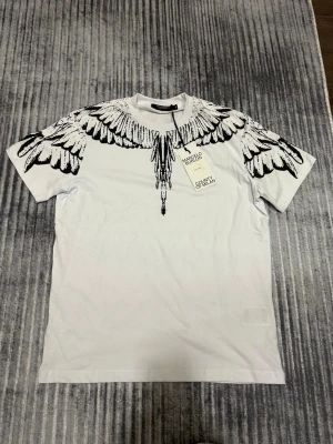 Vit t-shirt med vingmönster från Marcelo Burlon - Säljer en vit t-shirt från Marcelo Burlon med ikoniskt svart vingmönster över axlar och bröst. T-shirten har rund hals och korta ärmar. Perfekt för dig som vill sticka ut med en unik och trendig design. Skicka gärna prisförslag, va inte rädda och skäms inte för att pruta. 
